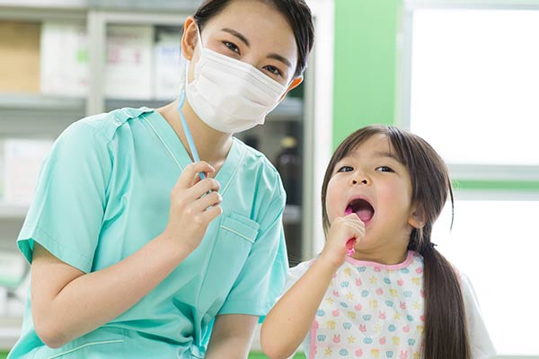 マスクを着けた歯科スタッフが、エプロンを着けた小さな女の子の口の中を診ている様子